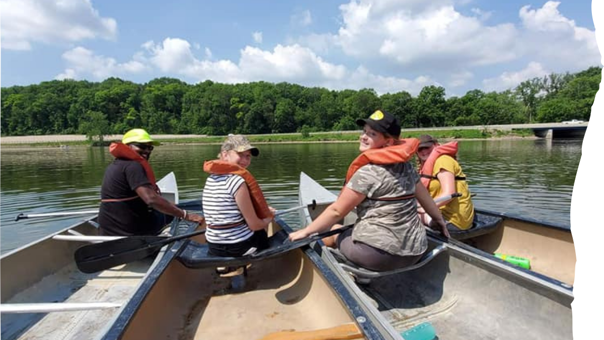 Four people rowing a boat on a lake with trees and a bridge in the background.