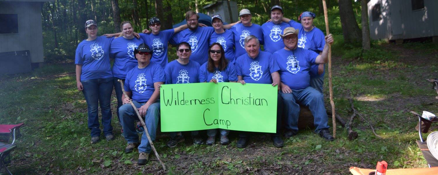 Group of people in blue shirts holding a sign for Wilderness Christian Camp outdoors.